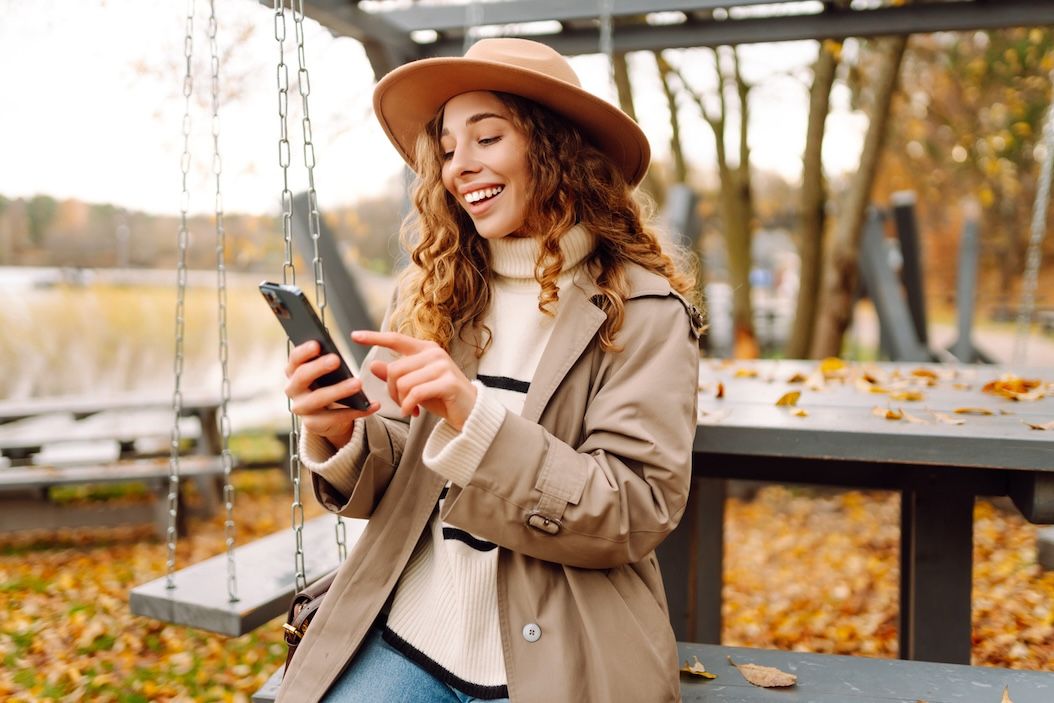 Young woman enjoying a fall day at a park swing, surrounded by autumn leaves, smiling while looking at her phone.